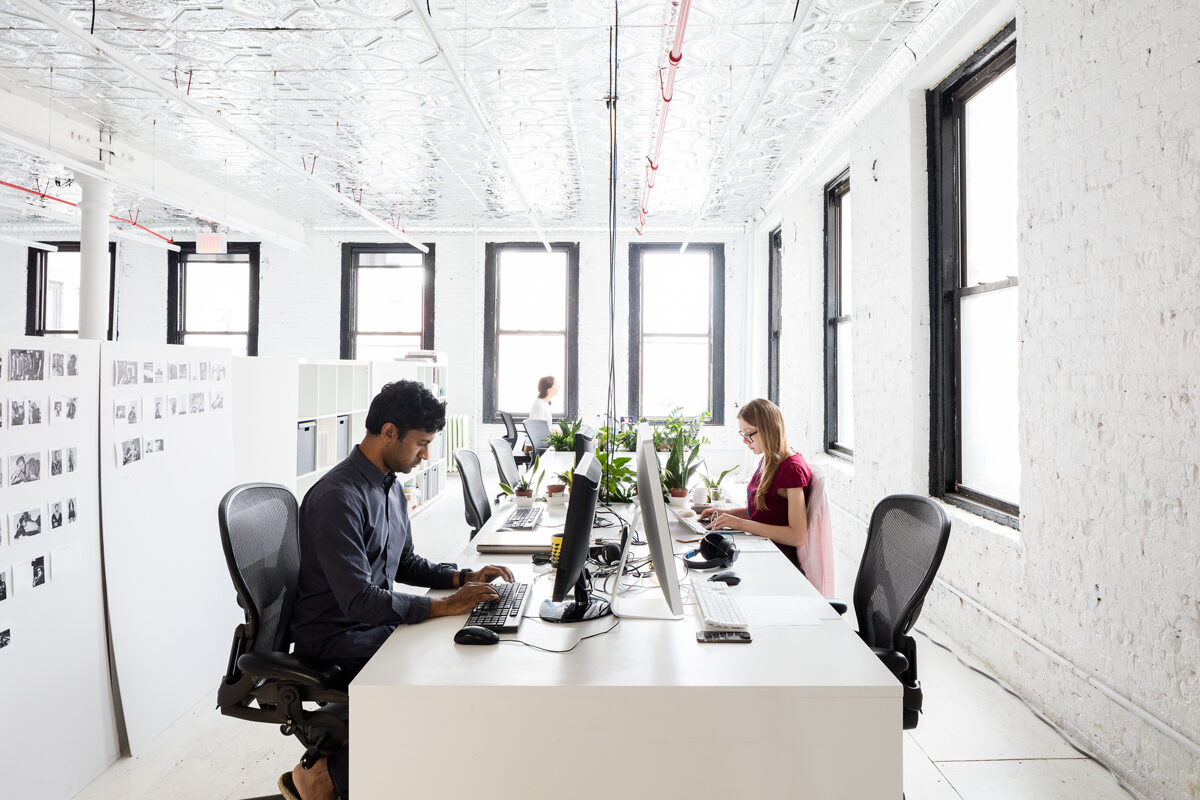 Members working at computers in a sunlit white-brick room with a tin ceiling