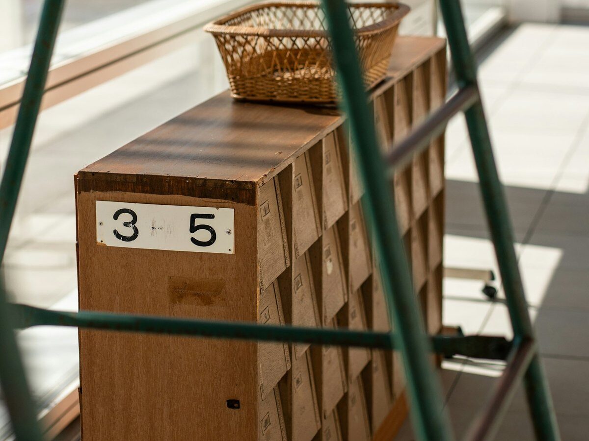 Sunlit wooden mail cubbies with a wicker basket on top, viewed through the frame of a green chair
